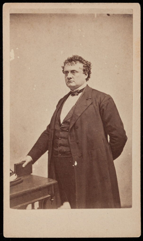 Studio portrait of a man photographed at a 3/4 angle from the waist up. His hair is curly and he wears a conservative suit. He is posing next to a table with a book in his right hand.